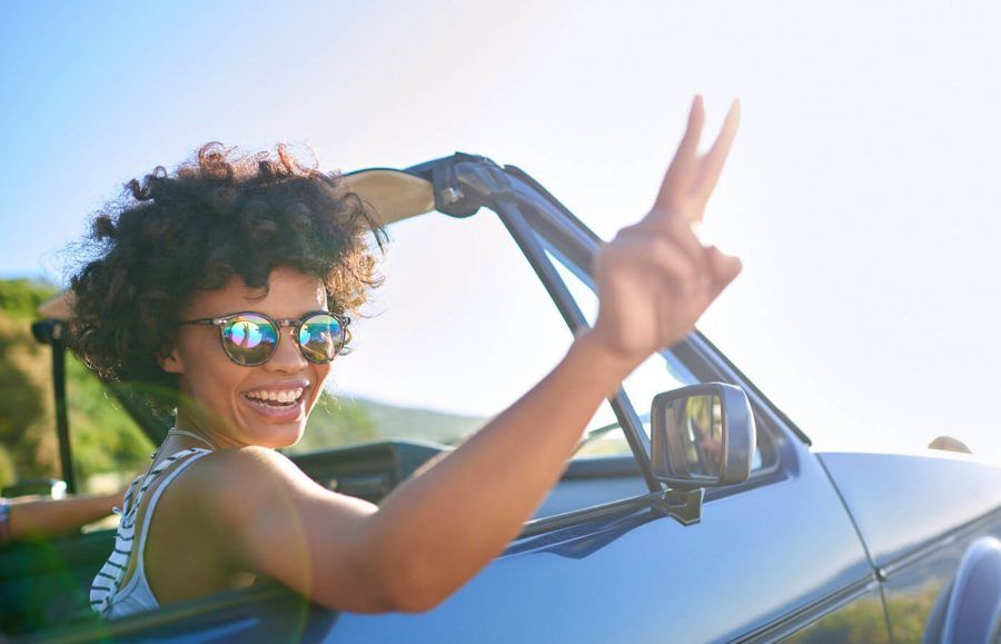 smiling woman in sunglasses holding up two fingers while riding in a convertible