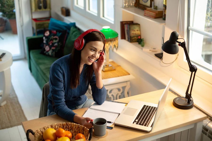 Attractive young woman teacher, teaching by holding an online class with her students from home using her laptop. Having a headphones on and smiling. Relaxed atmosphere and cozy ambient for the improvement of productivity.