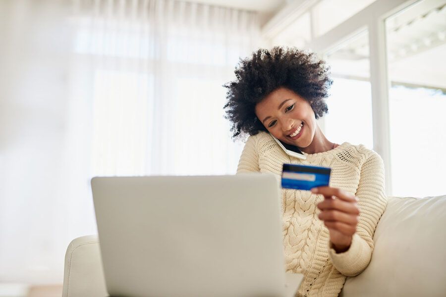 A woman wearing a white sweater sits on the couch as she smiles at her credit card and uses her laptop.