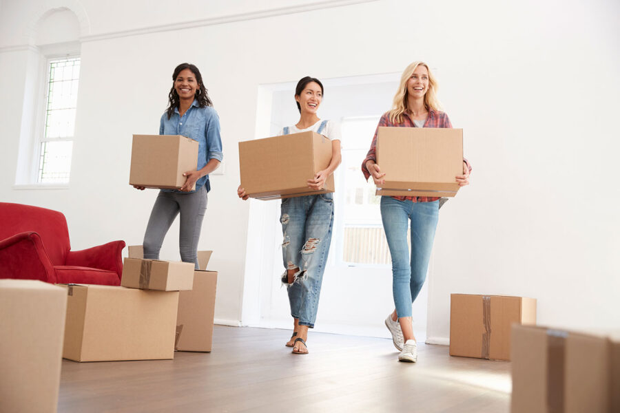 A group of three woman smile as they carry moving boxes into the living room of their new home.