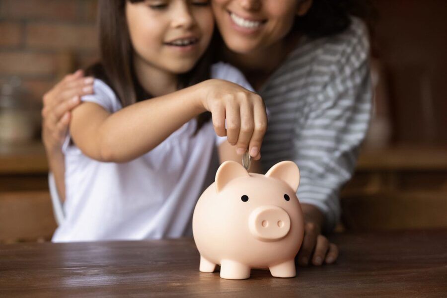 A mother embraces her daughter as she puts a coin in the piggy bank.