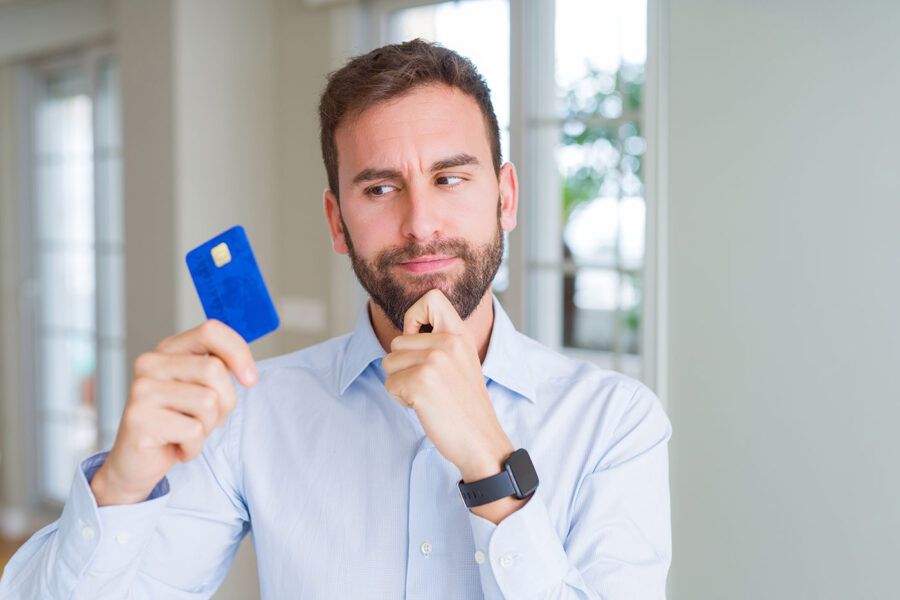 A man wearing a blue shirt has his hand to his chin as he looks at his blue credit card.