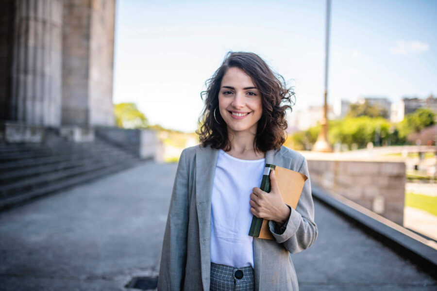 A law student smiles while holding a book outside a court building.