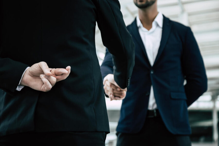 A man wearing a suit crosses his fingers behind his back as he shakes his other hand with another person.