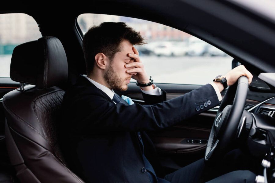 A man wearing a black suit has his hand to his face as he drives his car.