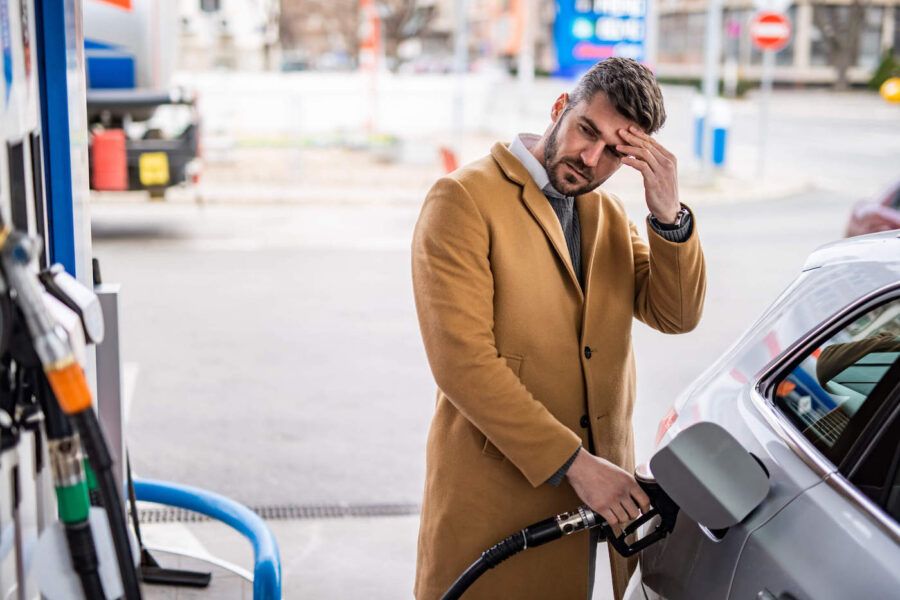 A man wearing a brown coat puts his hands to his head while looking at the gas pump at the gas station.