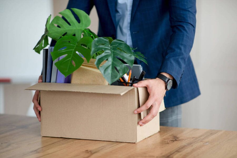 A man in a blue suit is holding a box that has a plant and his office supplies in it.