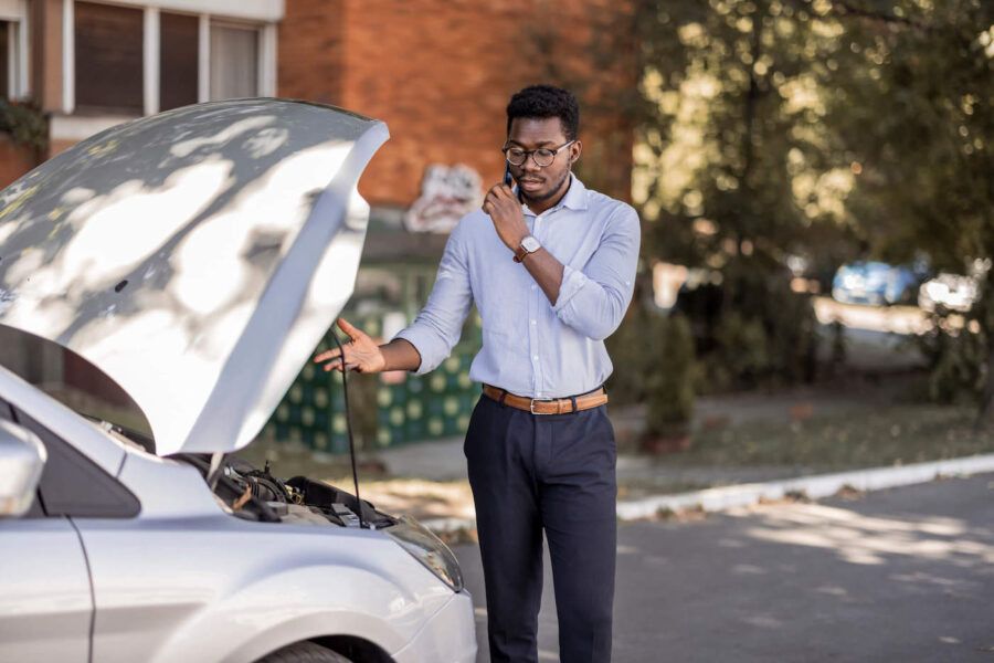 A man wearing a blue button up shirt, brown glasses, and gray pants is outside looking at his car's engine while on the phone.