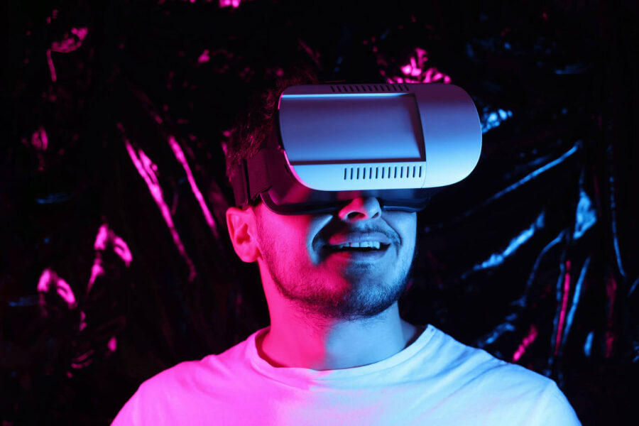 A man wearing a virtual reality headset is smiling as he is surrounded by pink and blue lighting.