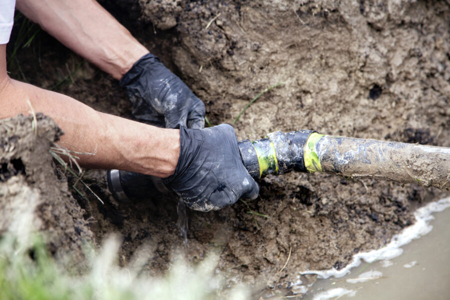 A man wearing black gloves repairs a sewer line that is surrounded by dirt.