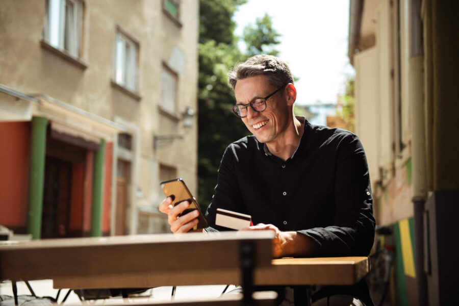 A man wearing a black shirt smiles at his phone and holds his credit card while sitting outside.