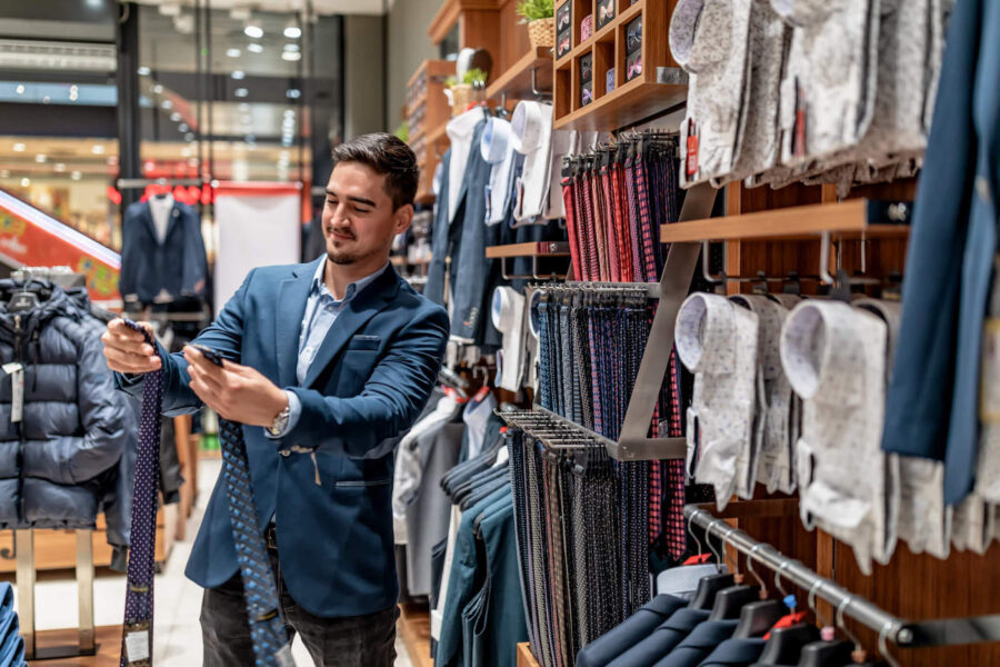 A man wearing a navy blue suit smiles while looking at ties inside a clothing store.