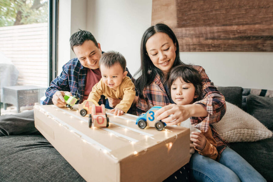 A mother and father are playing toy cars with their 2 young boys at their living room.