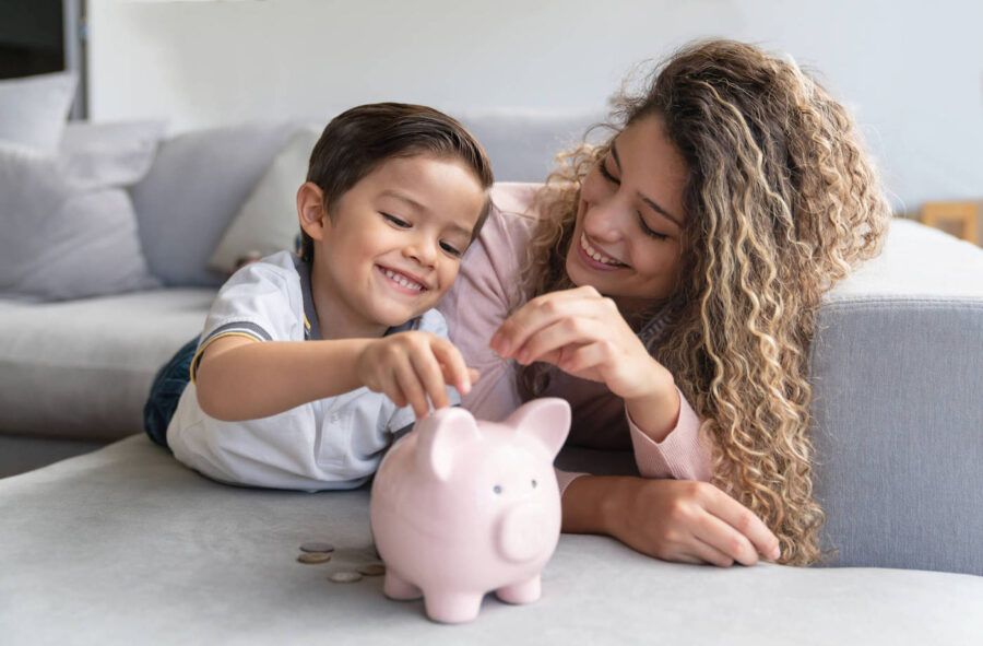 A mother and her son are laying down on the couch smiling as they put coins inside of a piggy bank.