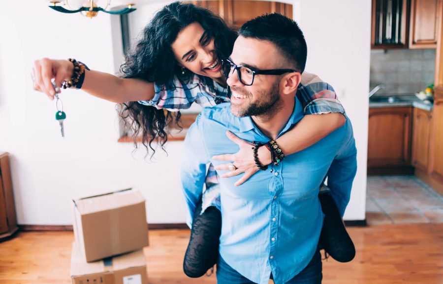 A woman being held up on a man's back, holding house keys out in front of her. A happy couple celebrating mortgage payments.