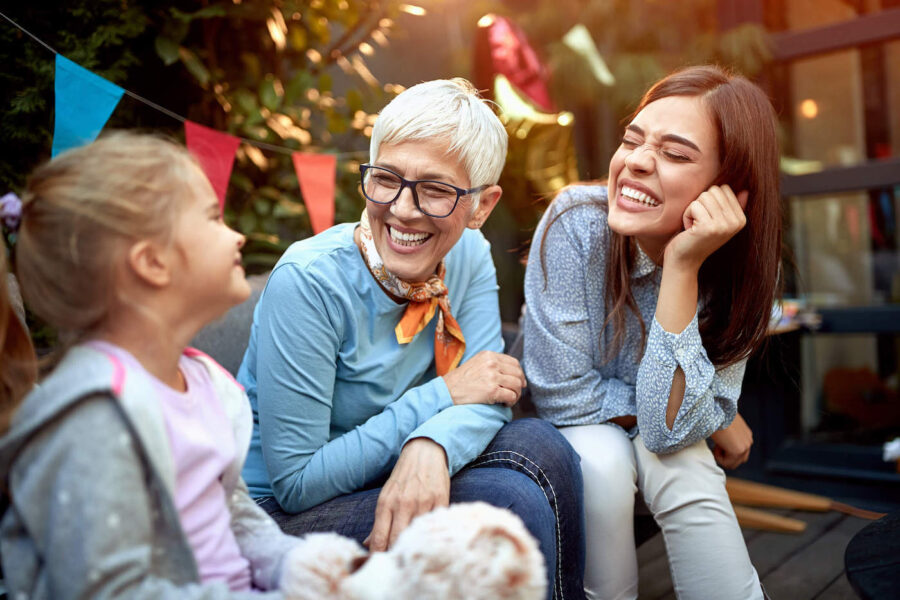 A mother, daughter and granddaughter are sitting on a couch outside and smiling during a birthday party.