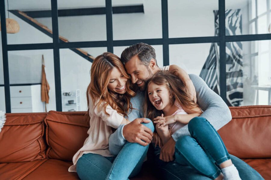Mother and father are hugging their daughter while laughing on the the couch.