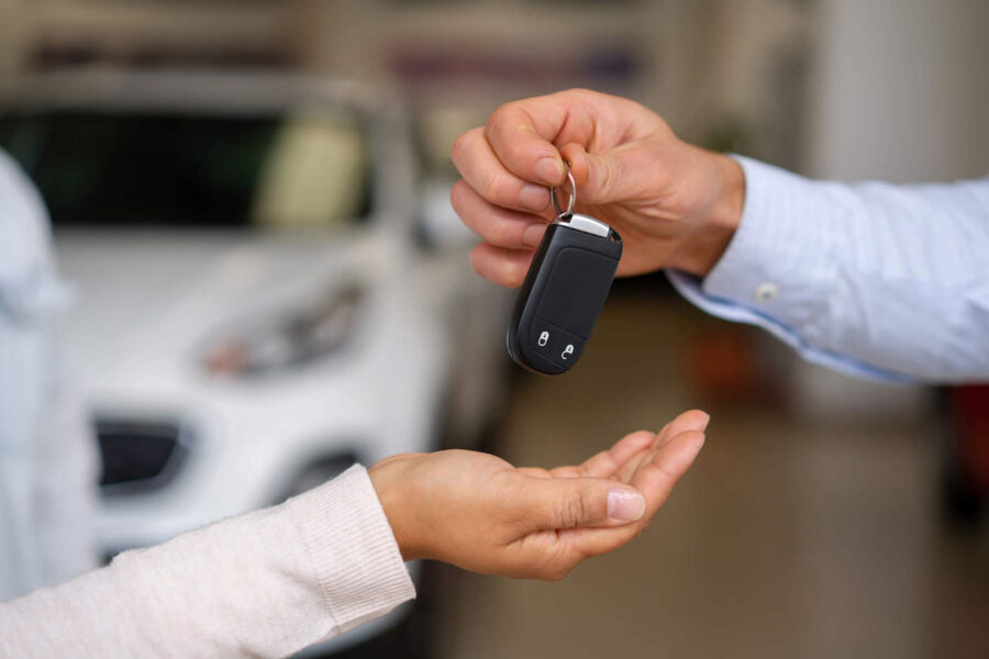 A hand holding car keys holds them above another hand with a car in the background.