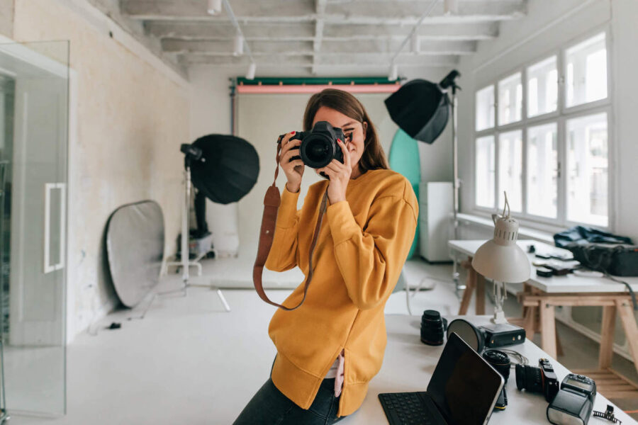 A photographer wearing an orange jacket uses her camera at her photography studio with equipment behind her.