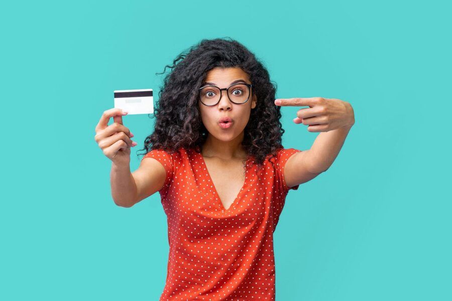 A woman has a surprised look on her face as she holds up her credit card and points at it with a light blue background behind her.