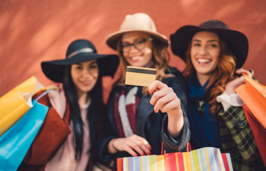 Three women wearing hats and holding shopping bags. The middle woman is holding a credit card up to the camera.