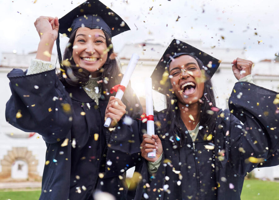 Two students celebrate graduating while holding their degrees while confetti falls from the sky.