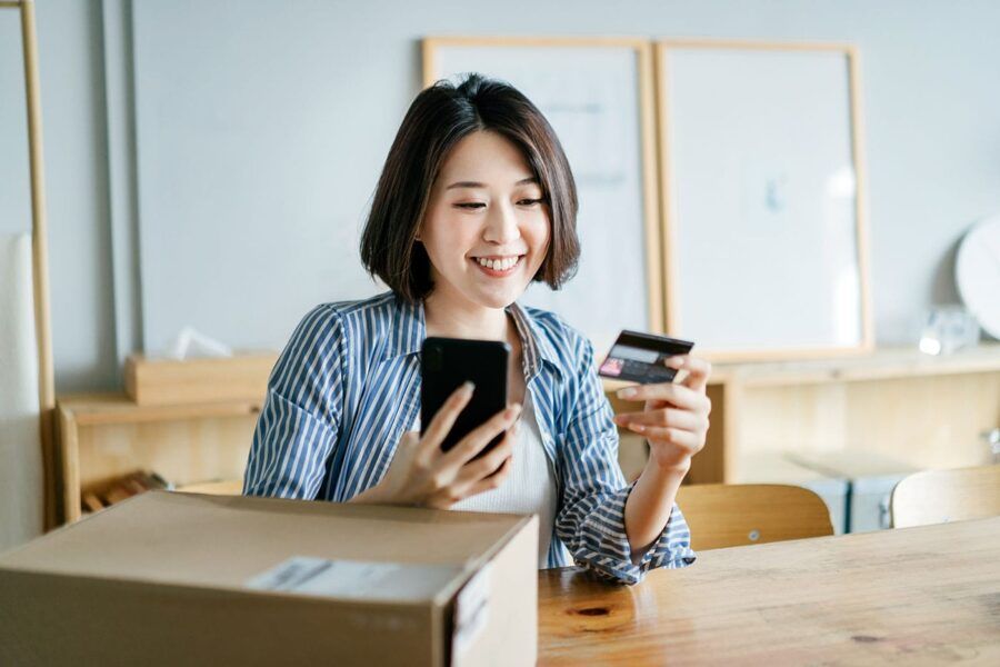 A woman wearing a striped blue shirt smiles at her credit card while holding her phone.