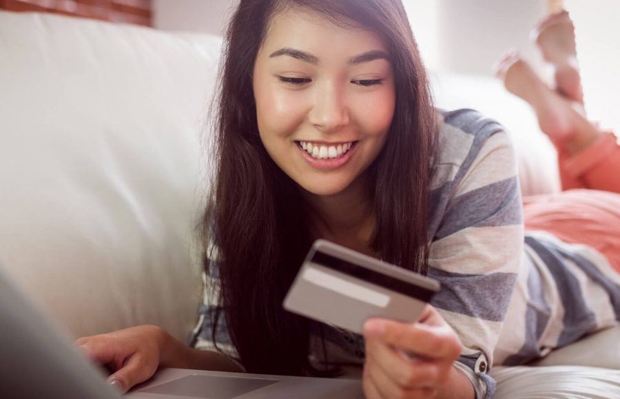 A young woman lays down on the couch while using her laptop and smiling at her credit card.