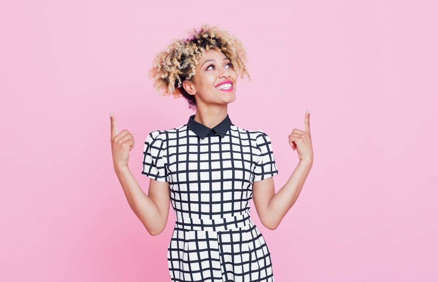 woman in black and white checkered dress pointing up with both hands against a pink background