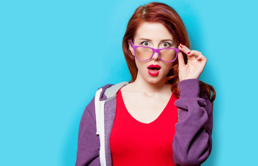 woman with red hair wearing a purple jacket and red shirt against blue background looking surprised while holding purple glasses
