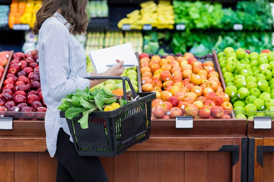 A woman looks at vegetables and fruits at the grocery store while holding a basket and small notepad.