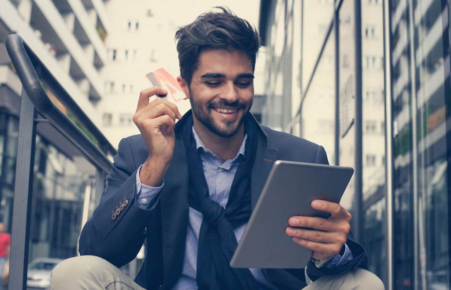 A man sits outside while holding his credit and smiling as he uses his tablet.