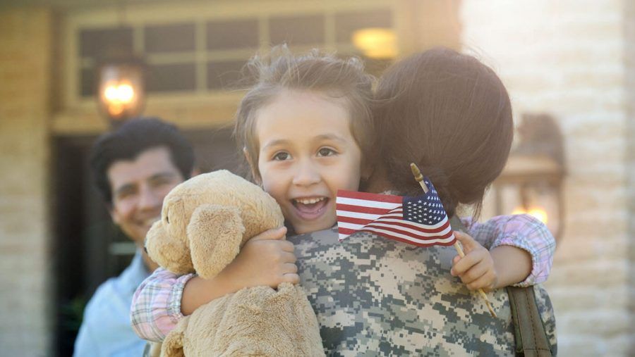 A child holding a plush animal and a small American flag, embracing a newly returned veteran family member.