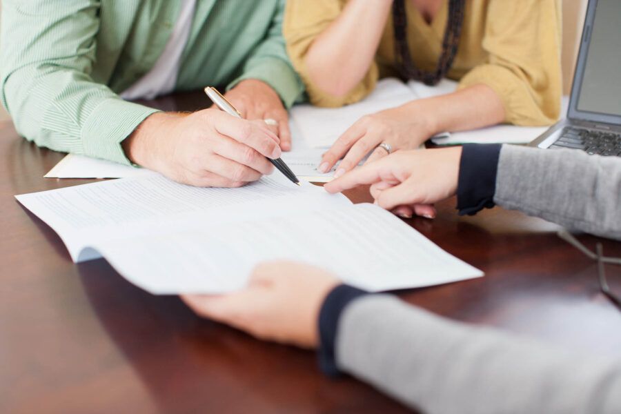 A couple and an agent look at documents while a man holds a pen to sign a document the agent is pointing at.