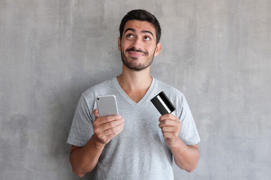 A man wearing a gray shirt looks up to the ceiling while he holds his phone in one hand and his credit card in the other.