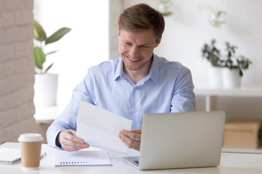 smiling man looking at paper seated a desk with an open laptop