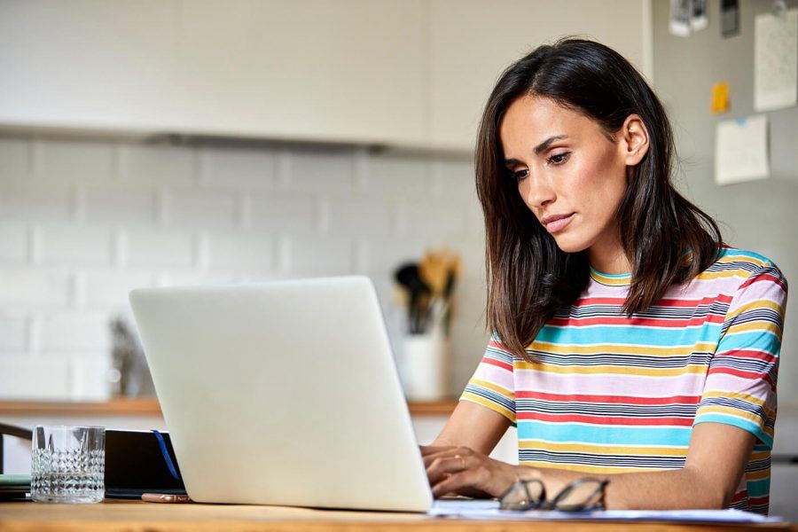 A woman wearing a colorful shirt uses her laptop at the kitchen table.