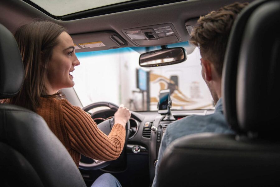 Woman wearing an orange sweater is driving while talking to a man in the passenger seat.
