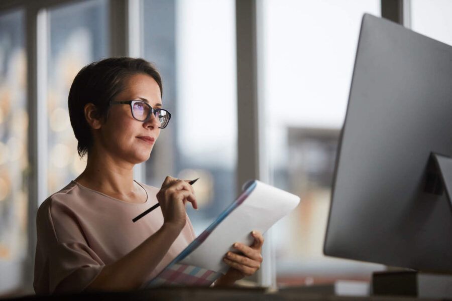 A woman in glasses takes notes on paper while looking at her computer.