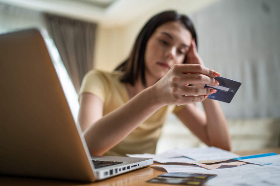 A woman is leaning on her hand while looking at her credit card with documents and an open laptop on the table.