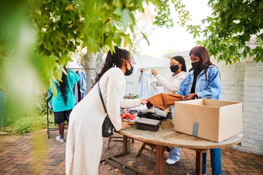A group of three woman look at clothes and items at a garage sale outside while wearing black masks.