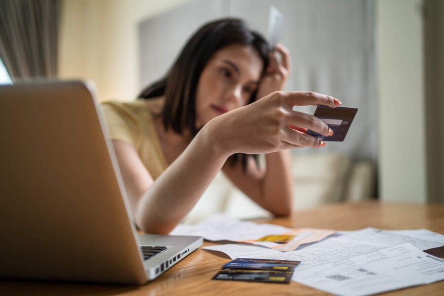 A woman leaning on her hand looks at her credit card while her laptop, other credit cards, and documents are on the table.