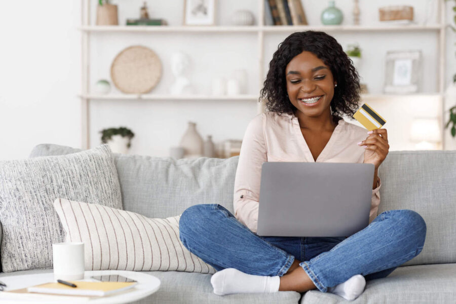 A woman sitting on her couch smiles at her laptop while holding up her credit card.