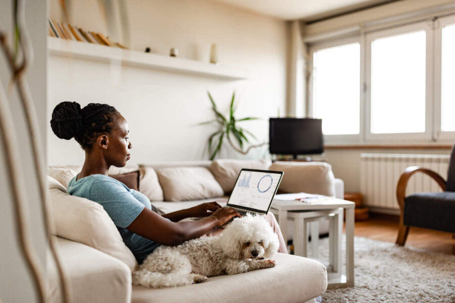 A woman sitting on the couch uses her laptop while her dog lays down next to her at home.