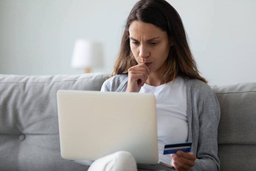 A women is sitting on the couch while looking at her laptop and holding her credit card.