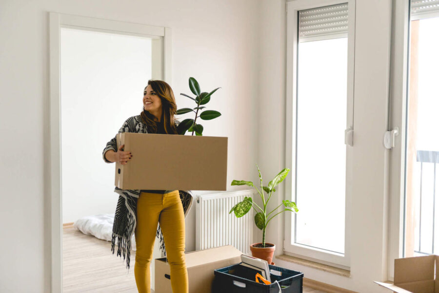 A woman smiles as she carries a box with a plant sticking out into her new home.