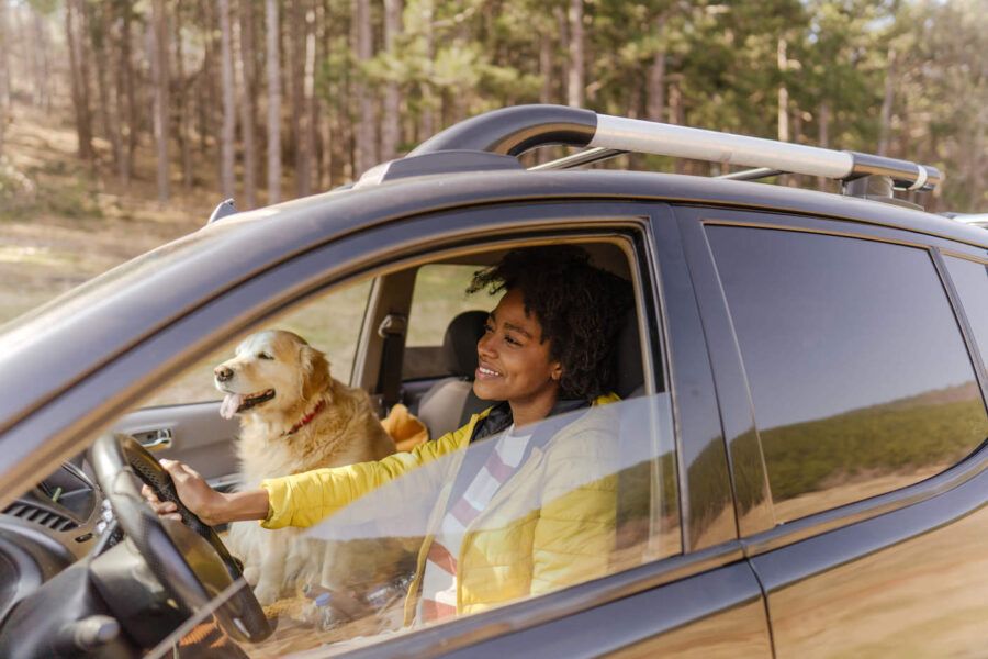 A woman wearing a yellow jacket smiles while driving with her dog in the passenger seat.