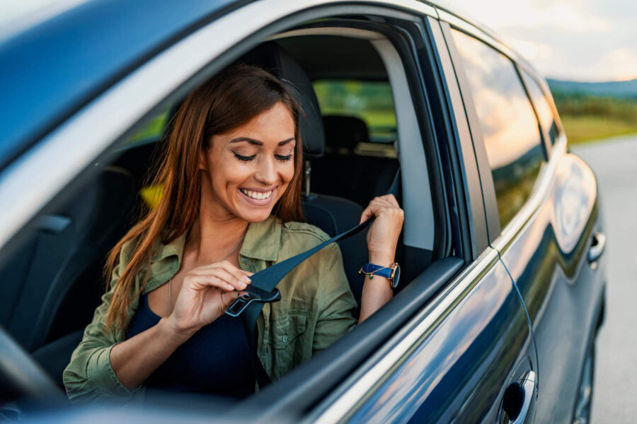 A woman smiles as she puts on her seatbelt to drive a car.