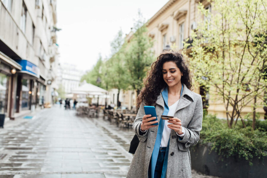 A woman wearing a gray coat smiles at her credit card while holding her phone outside.