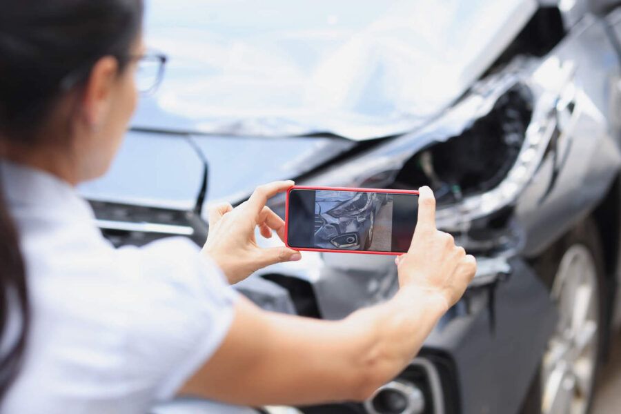 A woman holds up her red phone to take a picture of her damaged gray car.